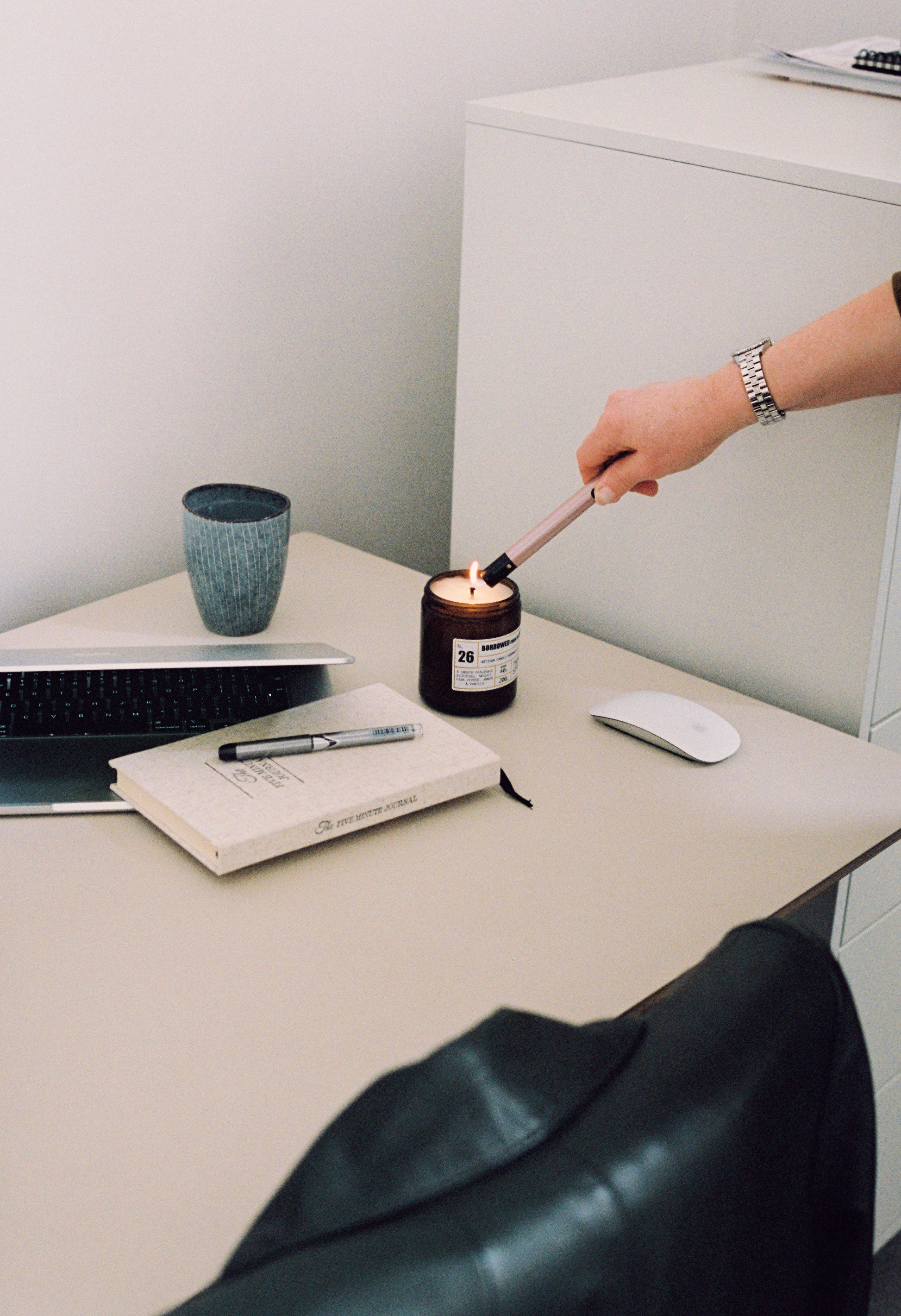 Person lighting a Thomas Street candle on a desk with a laptop, notebook, and mouse.